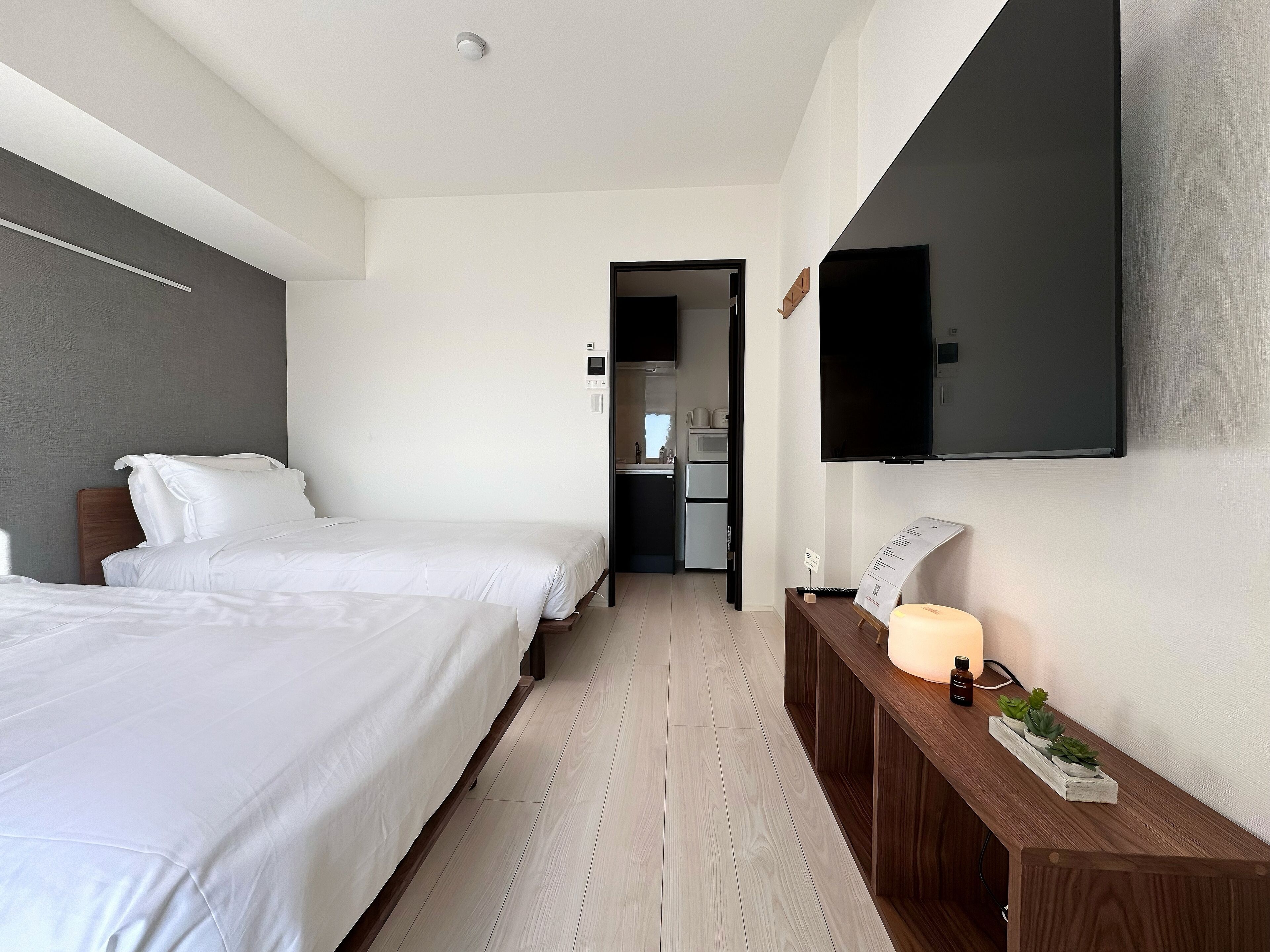 Habitación de hotel con dos camas, un televisor montado en la pared, una mesita de noche de madera con una lámpara y una planta pequeña.
