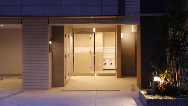 A building entrance with a tiled facade, a glass door, and a small garden area with plants and a lamp.
