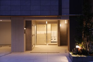 A building entrance with a tiled facade, a glass door, and a small garden area with plants and a lamp.