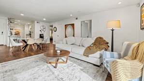 Una sala de estar moderna con un sofá blanco, una mesa de centro y un sillón a rayas. Hay un comedor con mesa y sillas, y una cocina al fondo.
