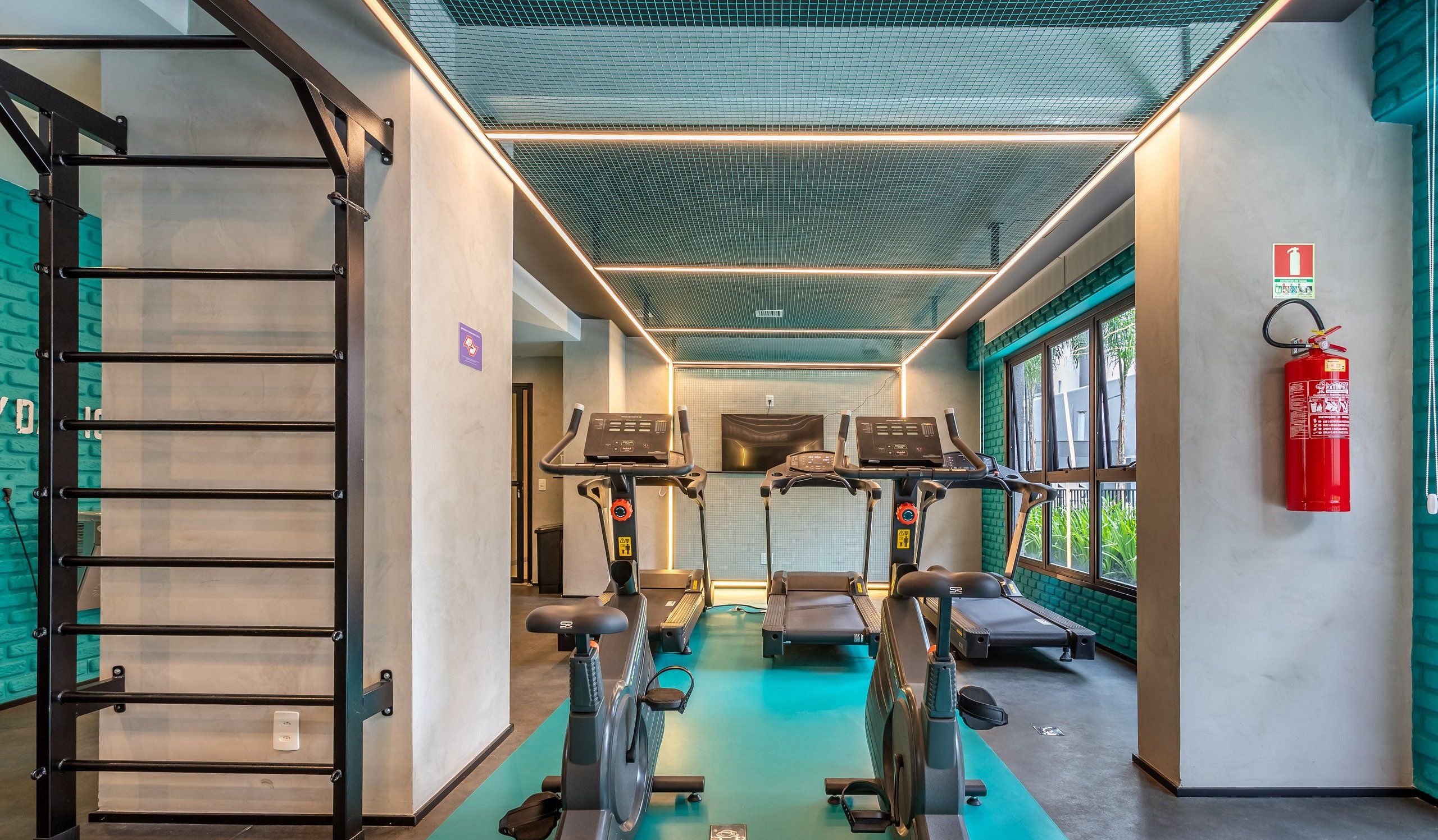 A gym with treadmills, stationary bikes, and a stair climber. There is a fire extinguisher on the wall and a window with a view of greenery.