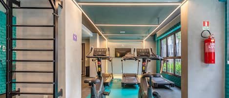A gym with treadmills, stationary bikes, and a stair climber. There is a fire extinguisher on the wall and a window with a view of greenery.