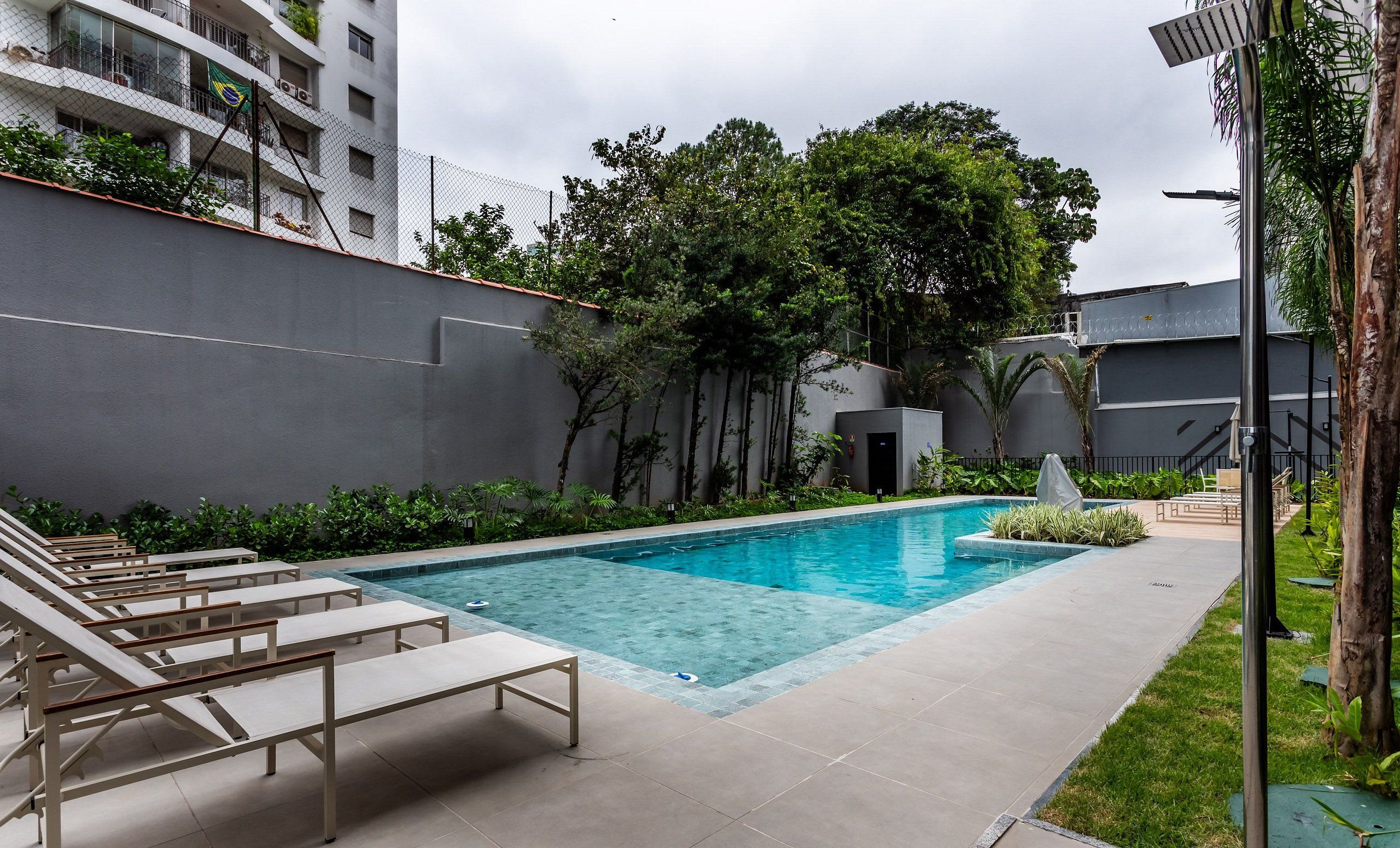 A swimming pool with lounge chairs, surrounded by greenery and a grey building.