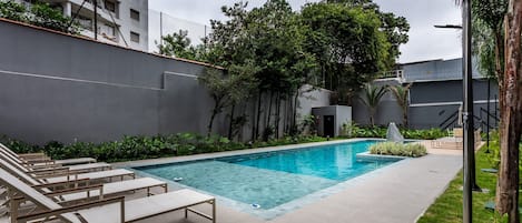 Piscina con sillones de descanso, rodeada de follaje y un edificio gris.