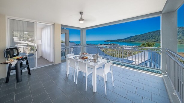 Un balcón con una mesa y sillas de comedor, una pequeña mesa con comida, y vistas al mar y las colinas.