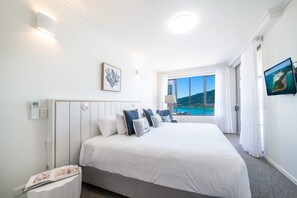 Ein Hotelzimmer mit einem großen Bett, einem Nachttisch, einem Fenster mit Meerblick und einem an der Wand befestigten Fernseher.