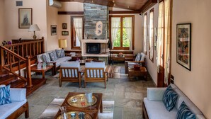 A spacious living room with a stone fireplace, wooden furniture, and large windows. - Villa Annelies Pelion (Volos)