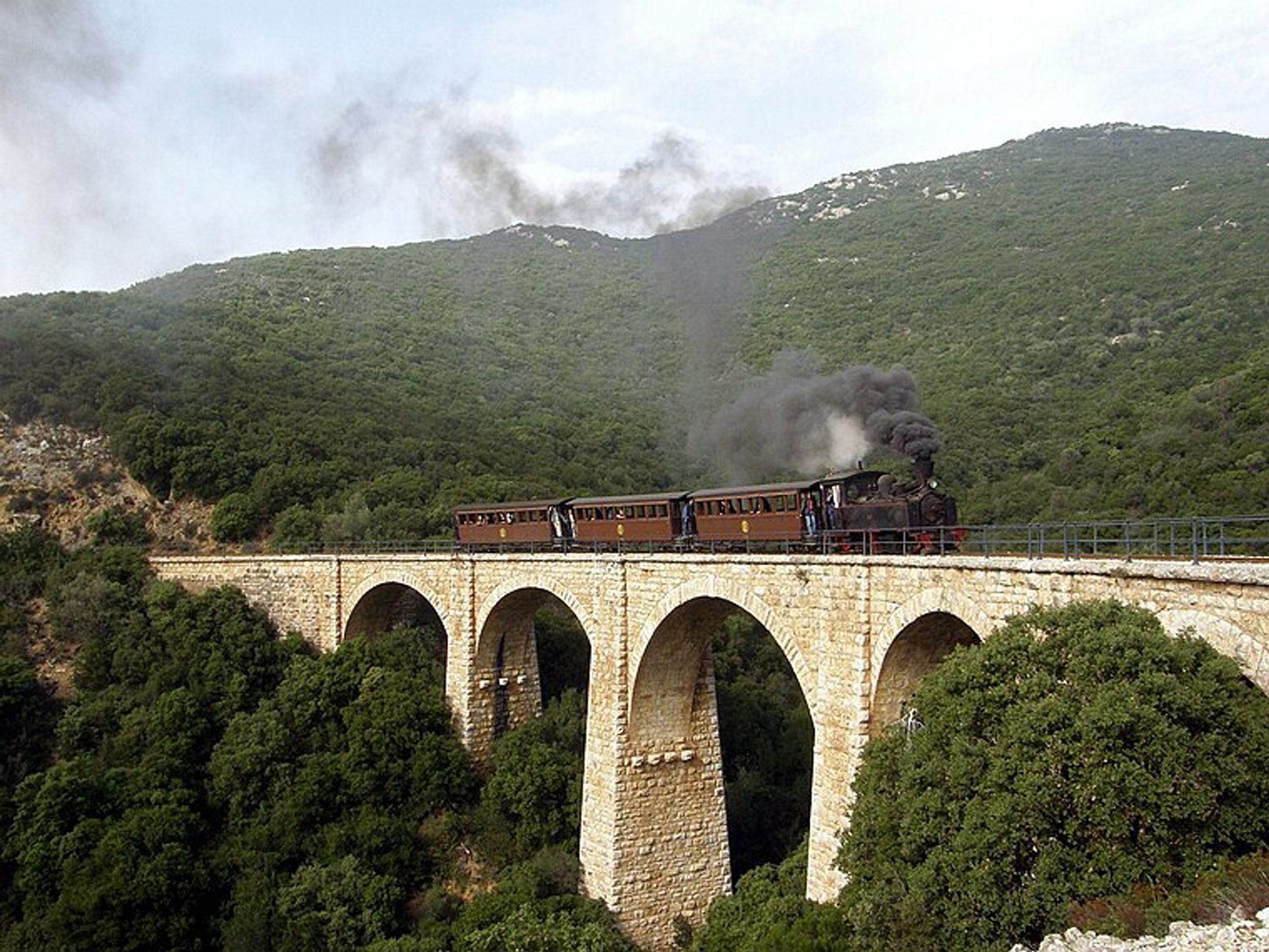A steam train crossing a stone bridge with multiple arches, surrounded by lush green hills.