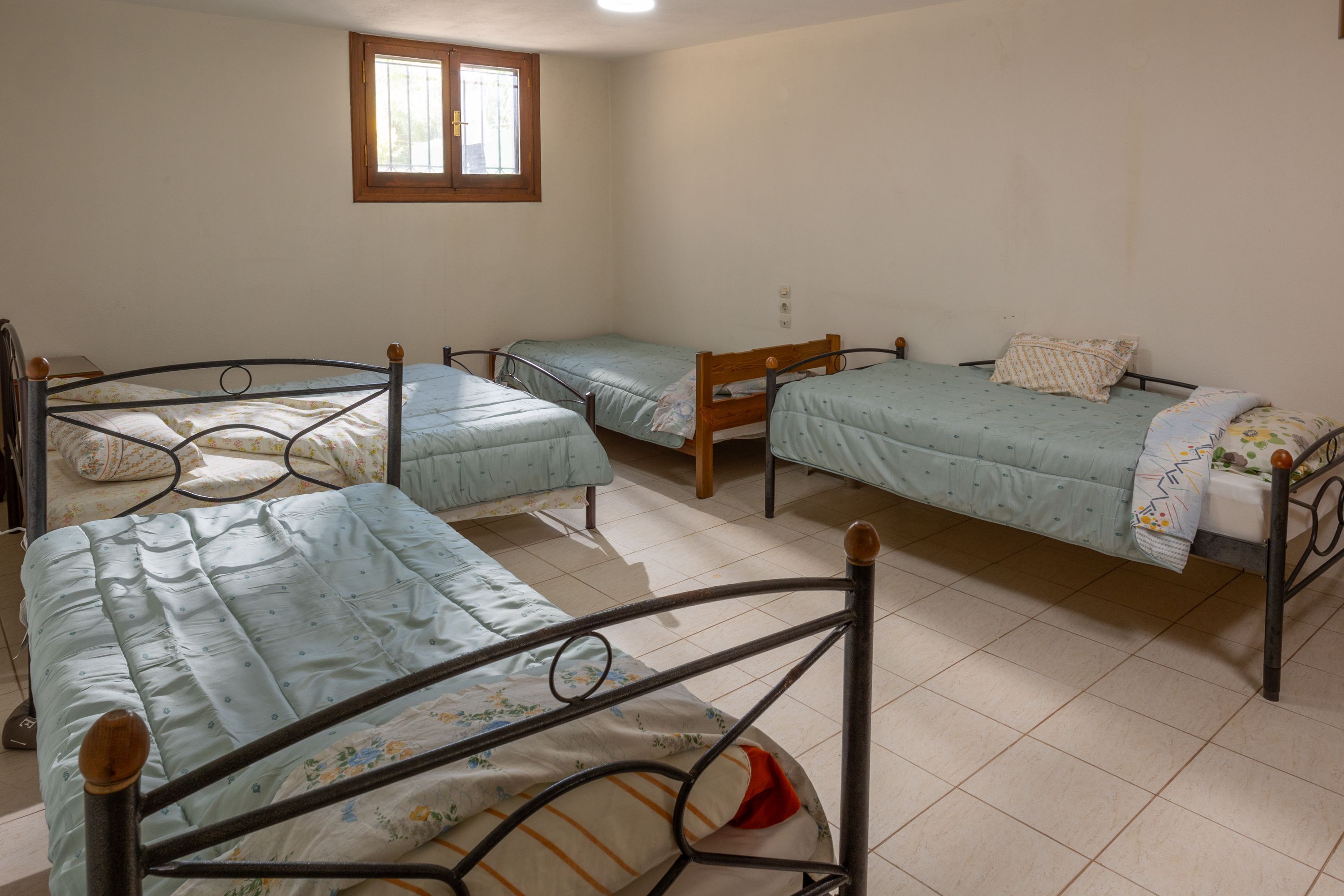 Ein Zimmer mit vier Betten, Metallrahmen, einem Fenster und Fliesenboden.