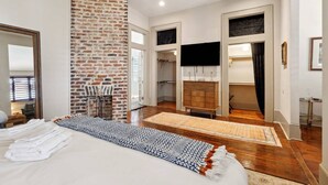 Une chambre avec un mur d’accent en briques, un lit avec du linge de lit blanc, une commode en bois et un téléviseur fixé au mur.