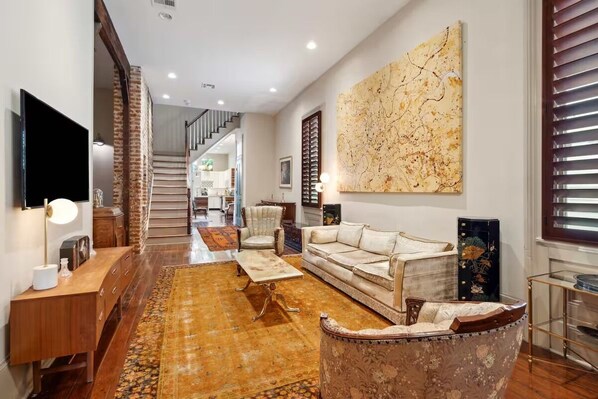 A living room with a sofa, armchair, and coffee table, a TV, and a large abstract painting on the wall. - 1 Block to Parade & St Charles, Heated Pool & Hot Tub, Walk to French Qtr (New Orleans)