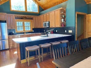 A kitchen with wooden cabinets, a large island with bar stools, a refrigerator, and a window with a view of trees.