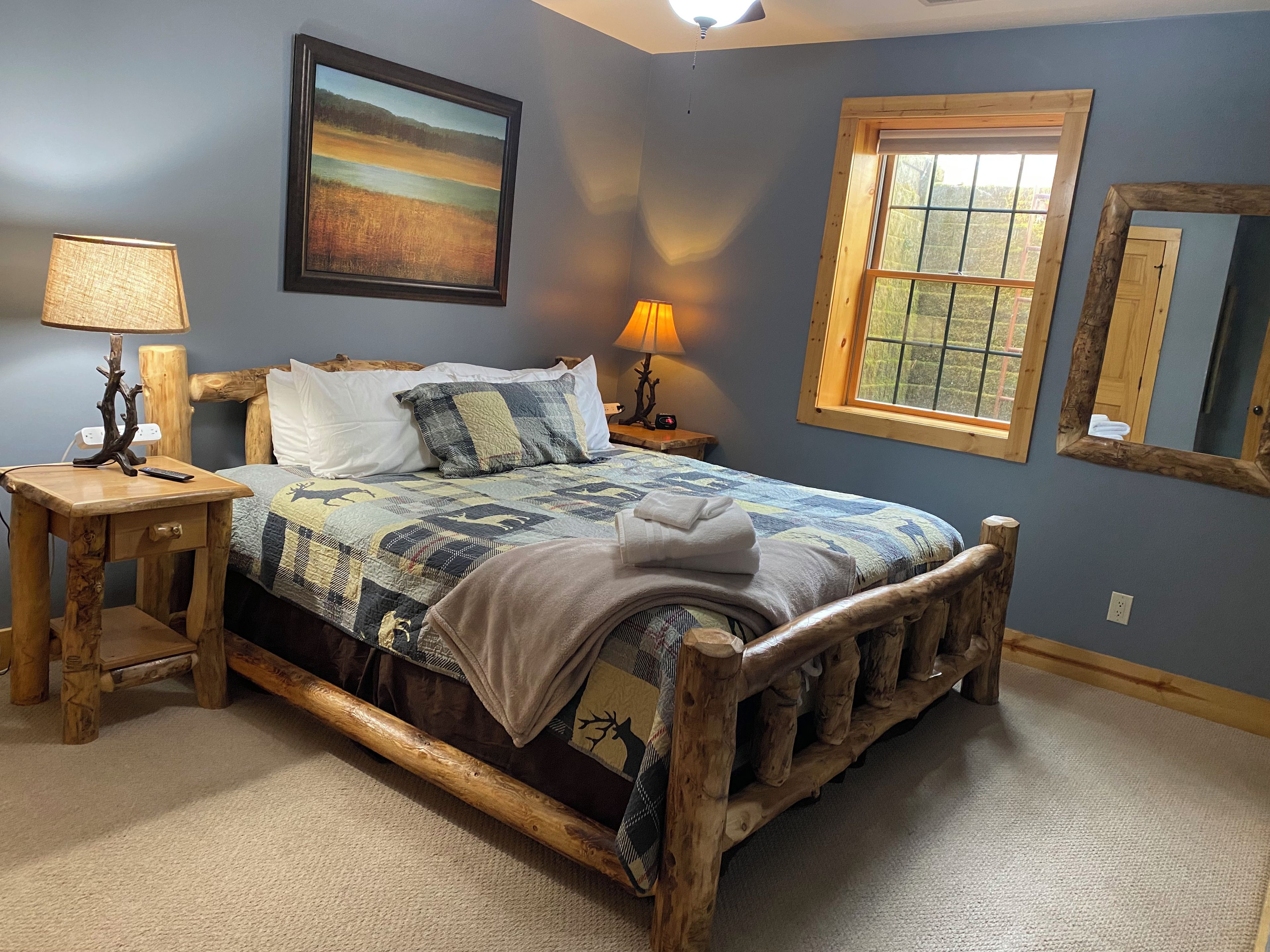 A bedroom with a wooden bed frame, two bedside tables with lamps, a window with a view, and a painting on the wall.