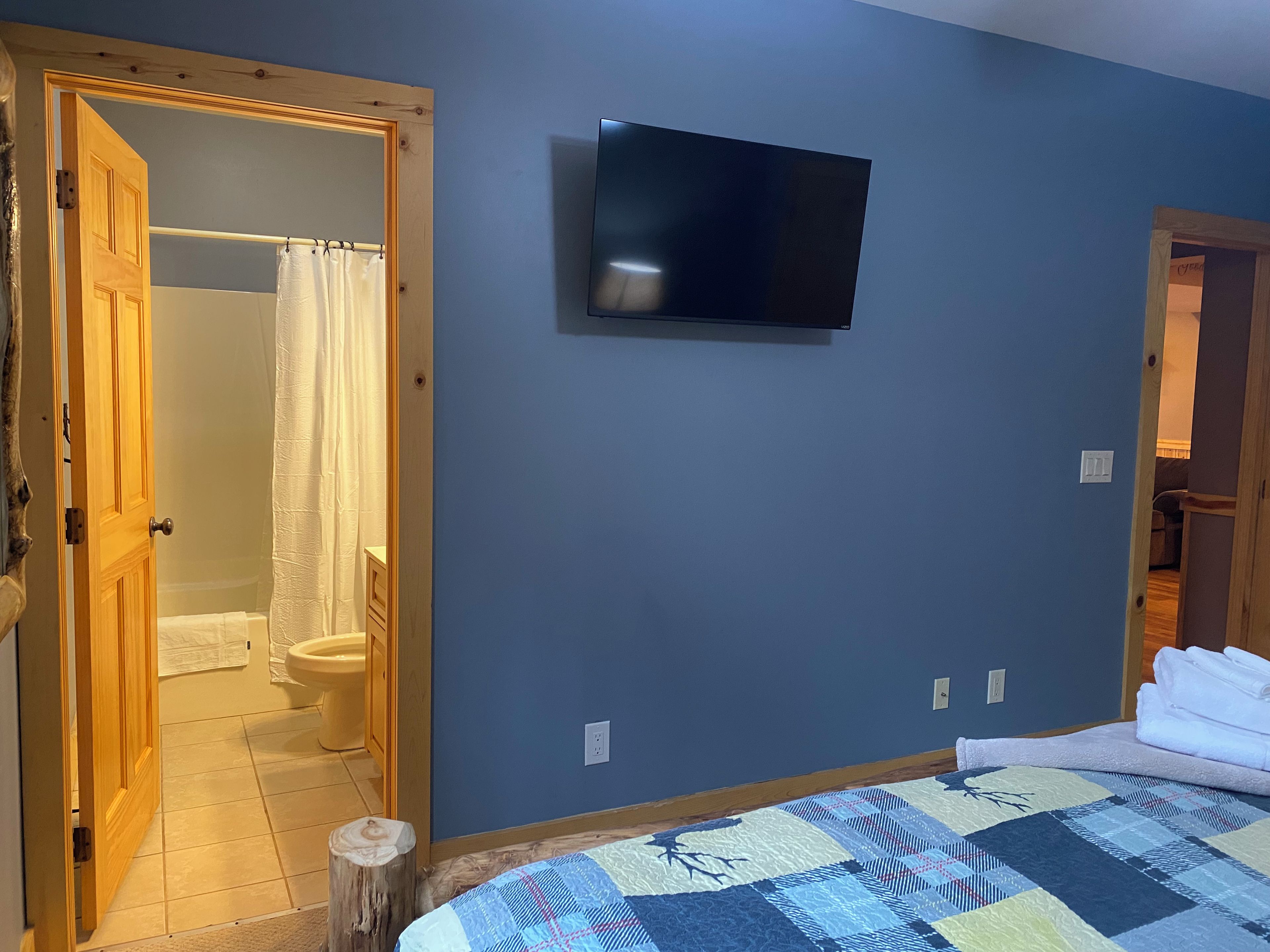 Un dormitorio con cama, televisor en la pared y baño visible a través de una puerta abierta.