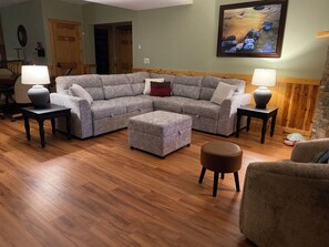 A living room with a sectional sofa, ottoman, two side tables with lamps, wooden flooring, and a painting on the wall.
