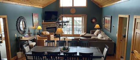 A cozy living room with a fireplace, a dining area with a wooden table and chairs, and a high ceiling with exposed beams.
