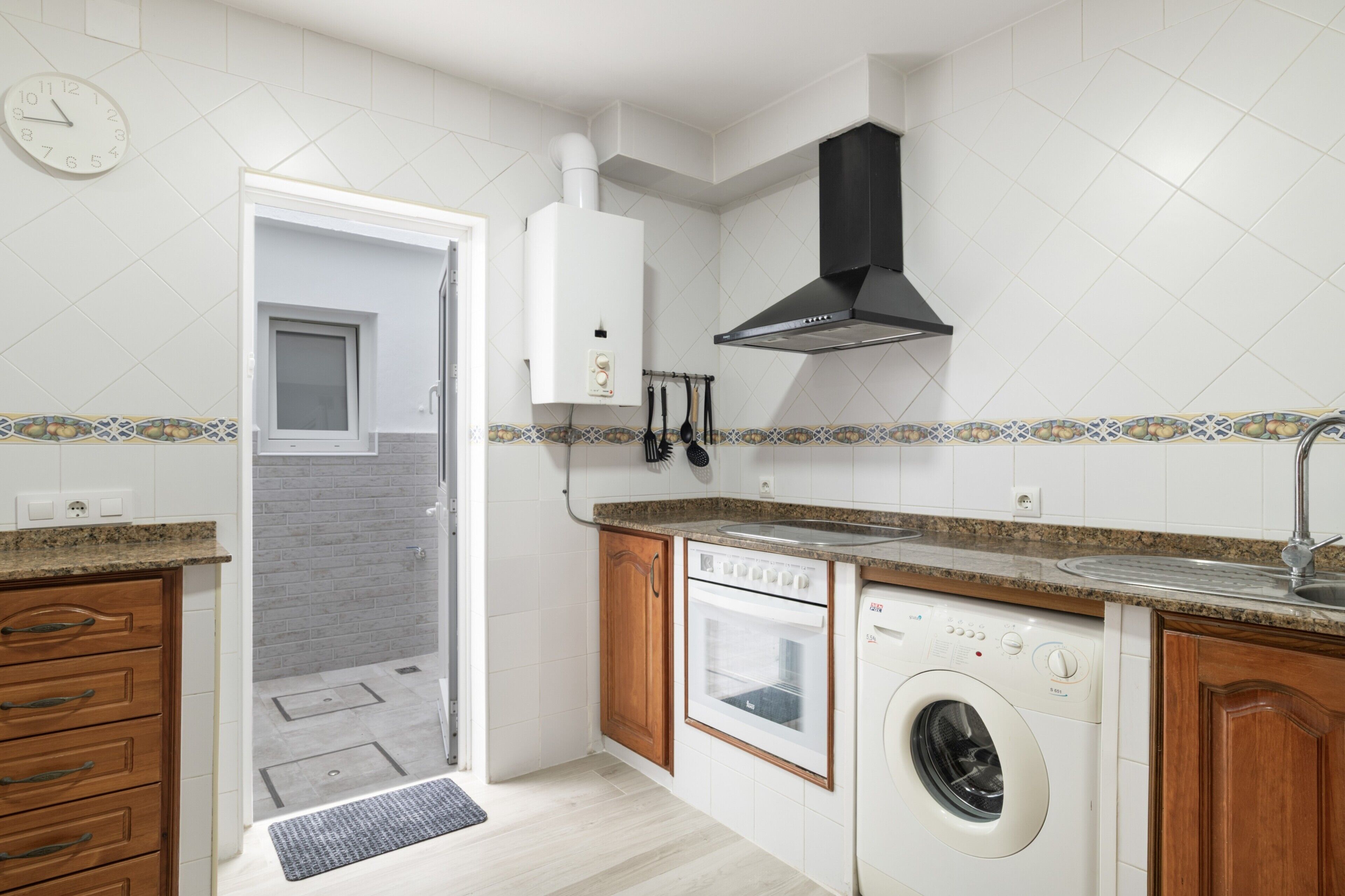 Une cuisine équipée d’un four encastré, d’un lave-vaisselle et d’un lave-linge, avec une fenêtre et une horloge au mur.