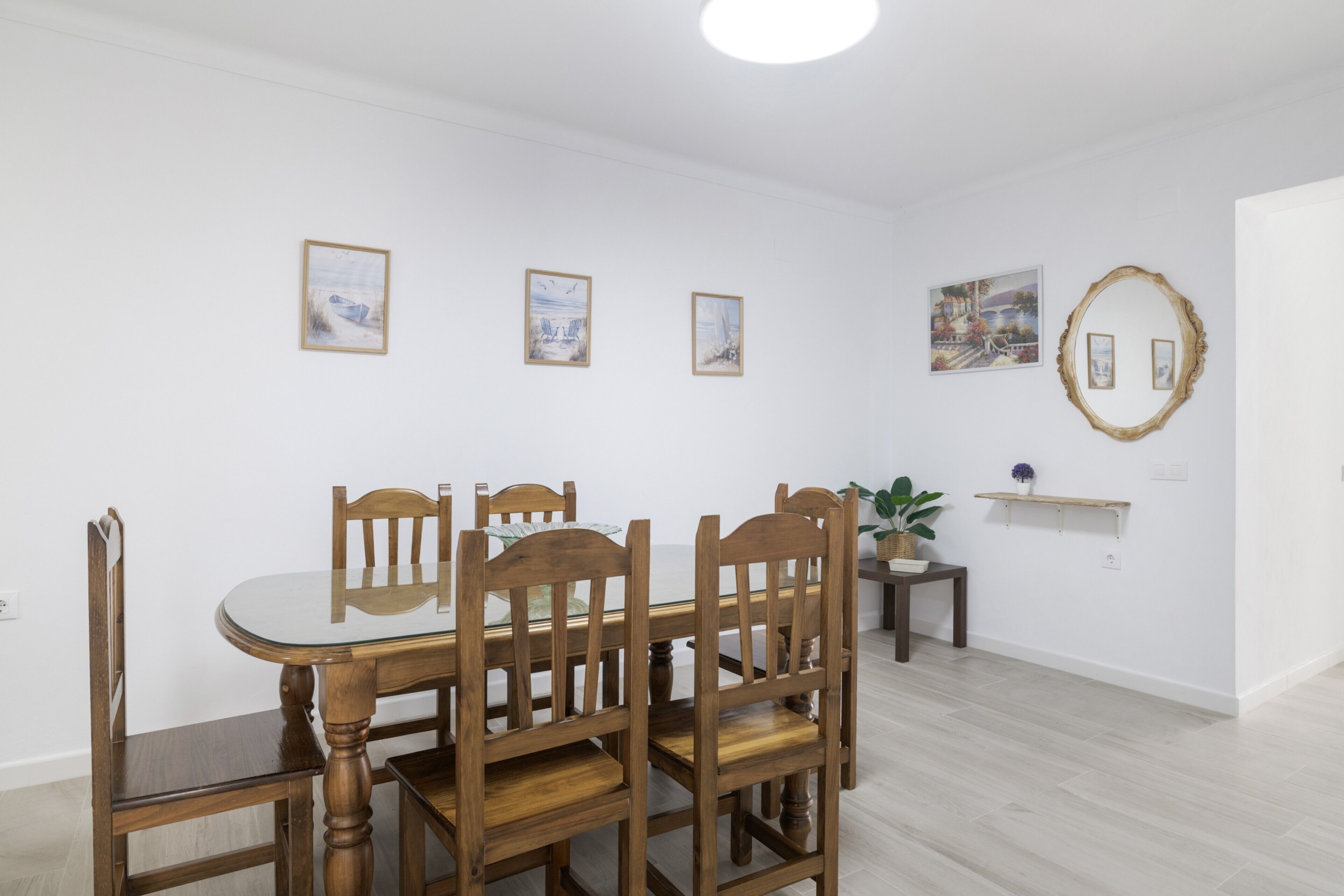 A dining area with a round glass table, wooden chairs, and a small side table with a plant.