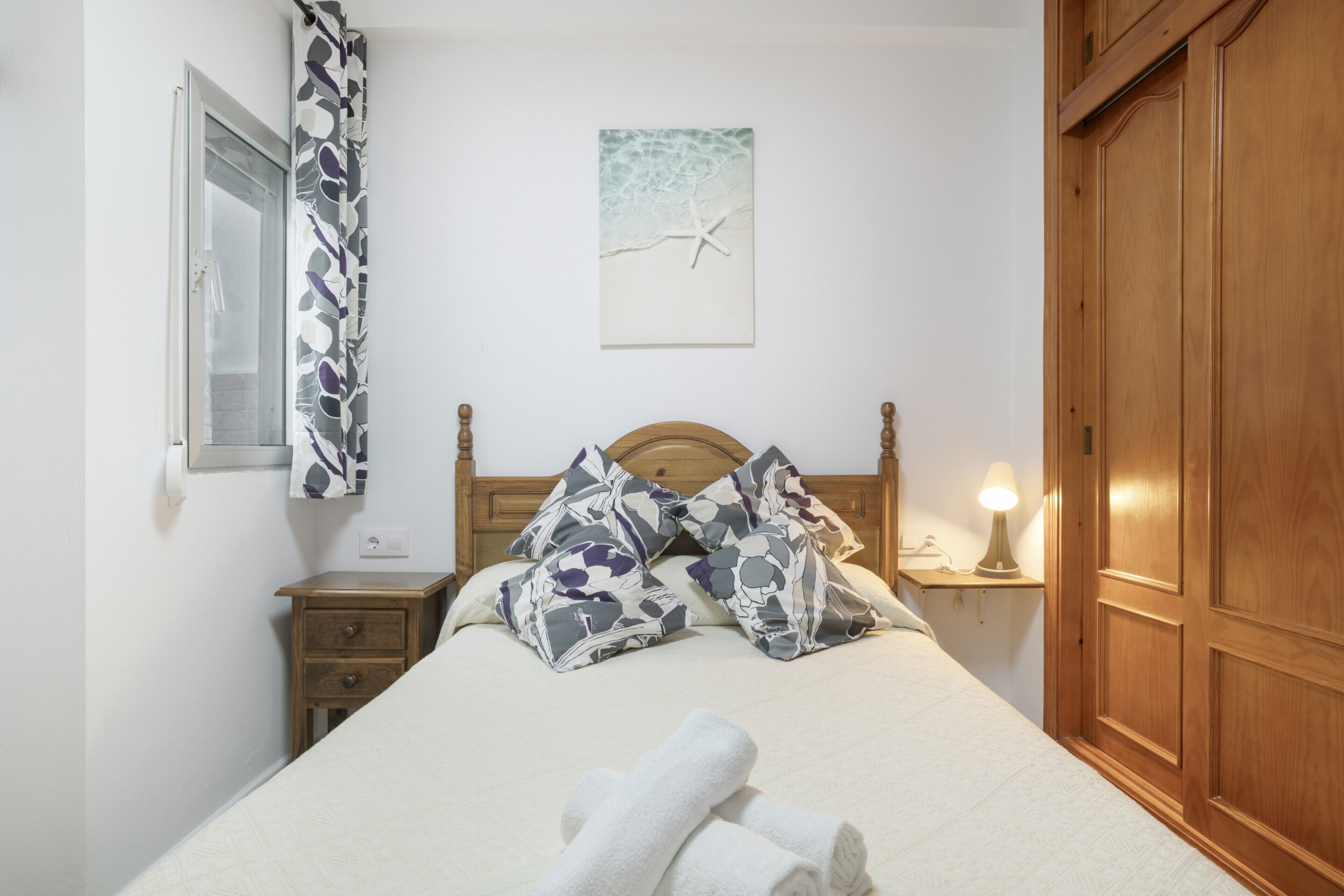 A bedroom with a bed, wooden headboard, a nightstand with a lamp, a wooden wardrobe, and a wall with a painting.
