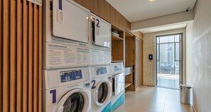 A laundry area with multiple washing machines, a trash bin, and a view of the outside through a glass door. - HMO - Full-length Studios in Building c/ Pool (São Paulo)