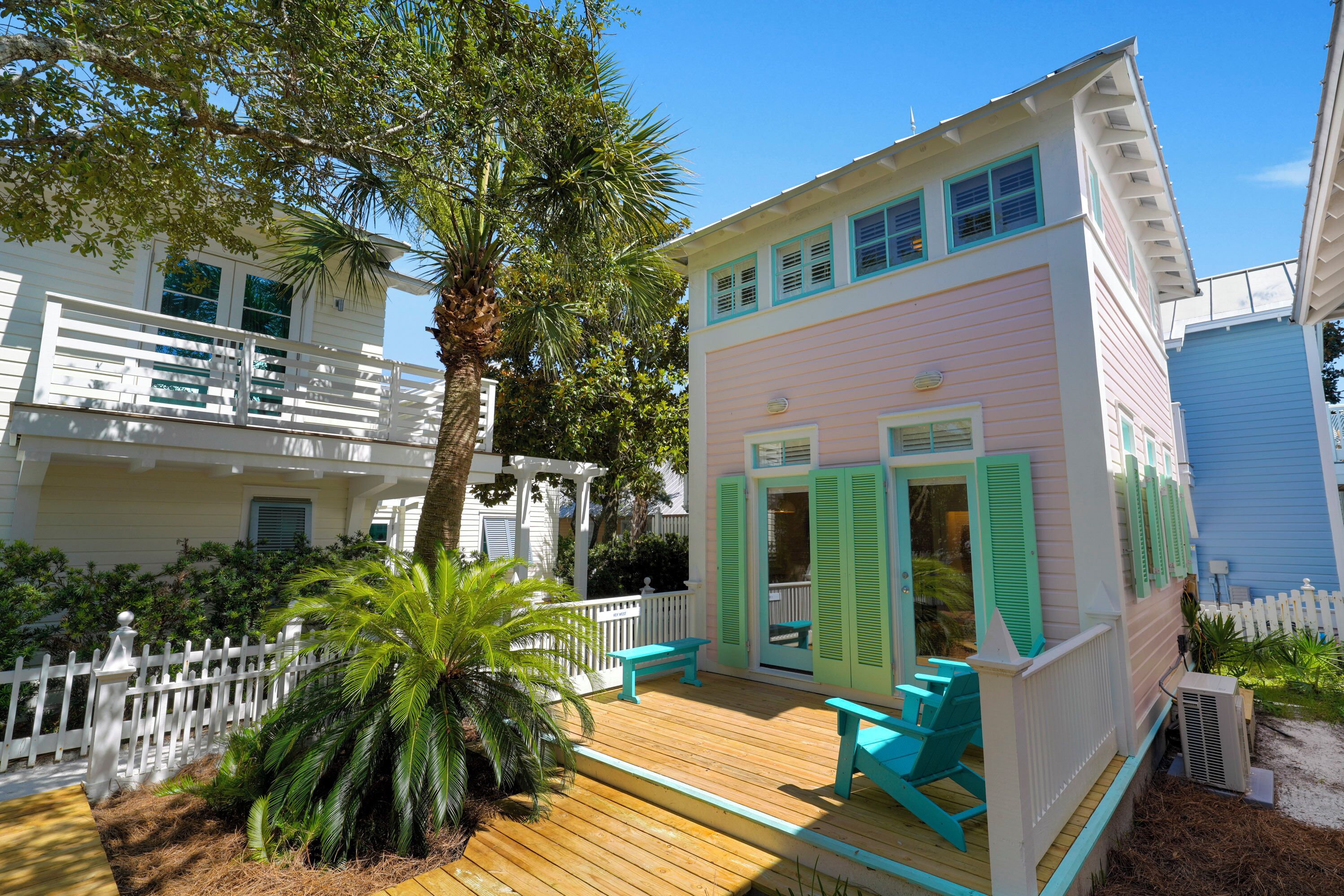 A two-story building with a balcony and a deck, surrounded by greenery and a white picket fence.