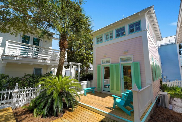 A two-story building with a balcony and a deck, surrounded by greenery and a white picket fence.