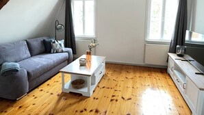 A modern living room with a grey sofa, a white coffee table, and a wooden floor. There are two windows with curtains, a lamp, and a cabinet. - Vacation apartment Minkwitz in the nursery house in Fürstlich Drehna (Luckau, OT Fürstlich Drehna)