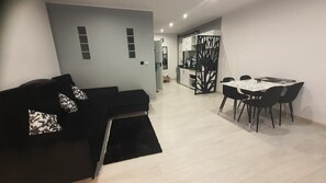 Un salon moderne avec un canapé noir, une table à manger avec des chaises, et une cuisine en arrière-plan.