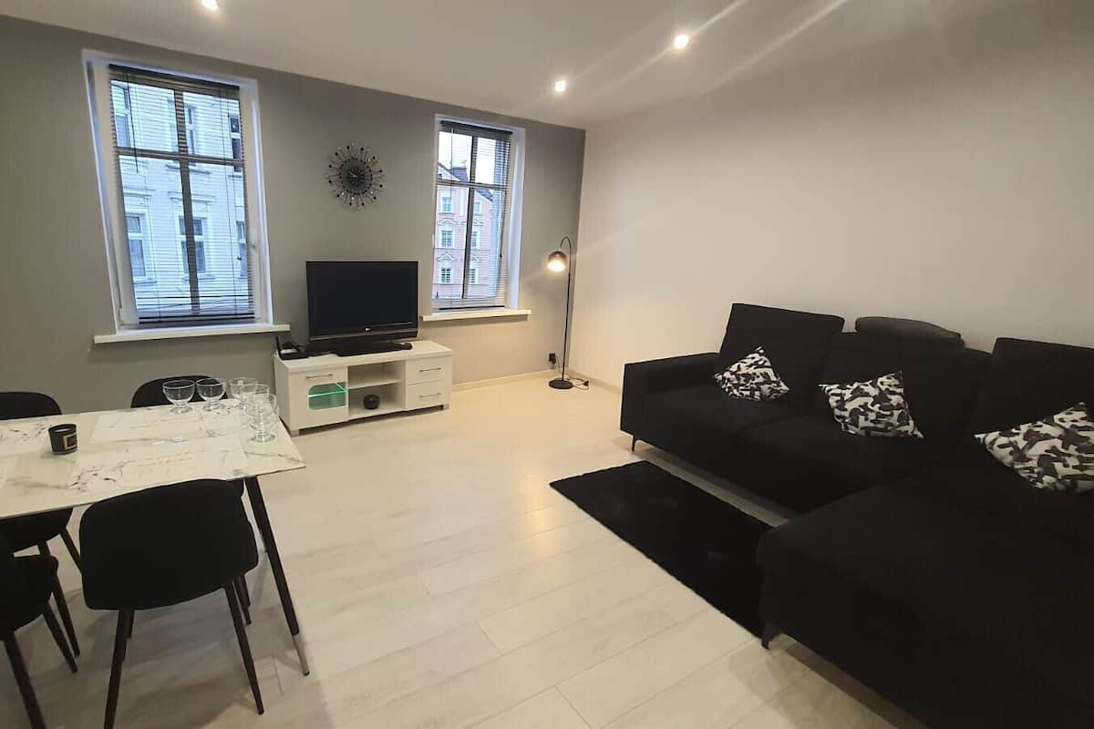A modern living room with a black sofa, a dining table with chairs, and a TV on a stand.