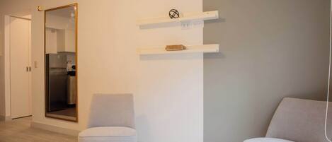 Un salon moderne avec deux fauteuils gris, une table basse blanche et un grand miroir au mur. On aperçoit des étagères blanches au-dessus des fauteuils, et un coin cuisine est visible au fond.