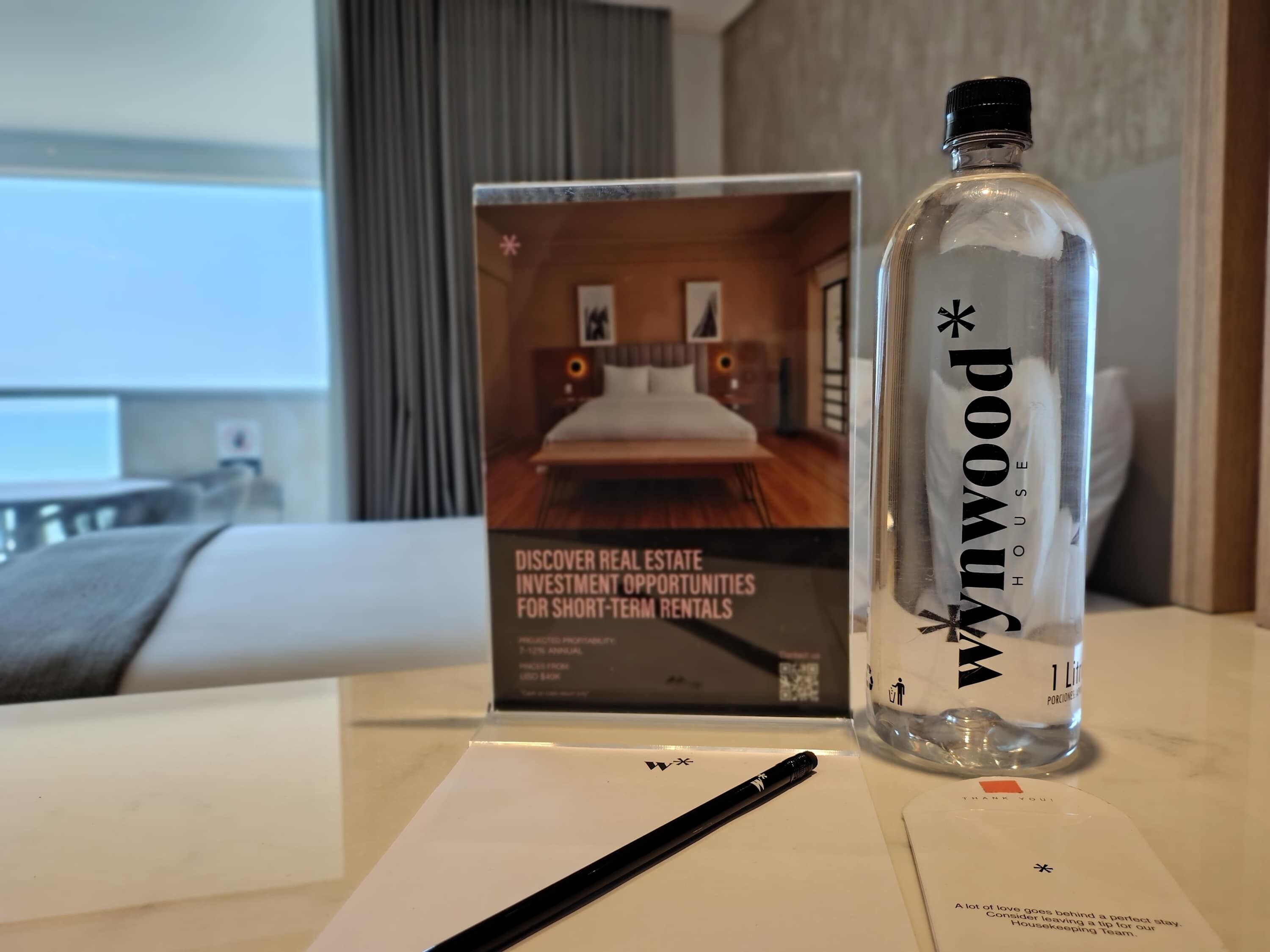 A hotel room with a bed, a bottle of water, a brochure, and a pen on the table.