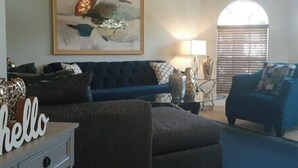 A living room with a blue sofa, a coffee table, and a lamp. There is a painting on the wall and a window with blinds.
