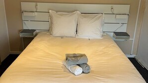 A neatly made bed with a quilted mattress, two pillows, and a rolled towel on the bed. There are bedside tables with lamps and a power outlet on the wall. - Mimosa room - Domaine de la Provençale (ST DIDIER DE LA TOUR)