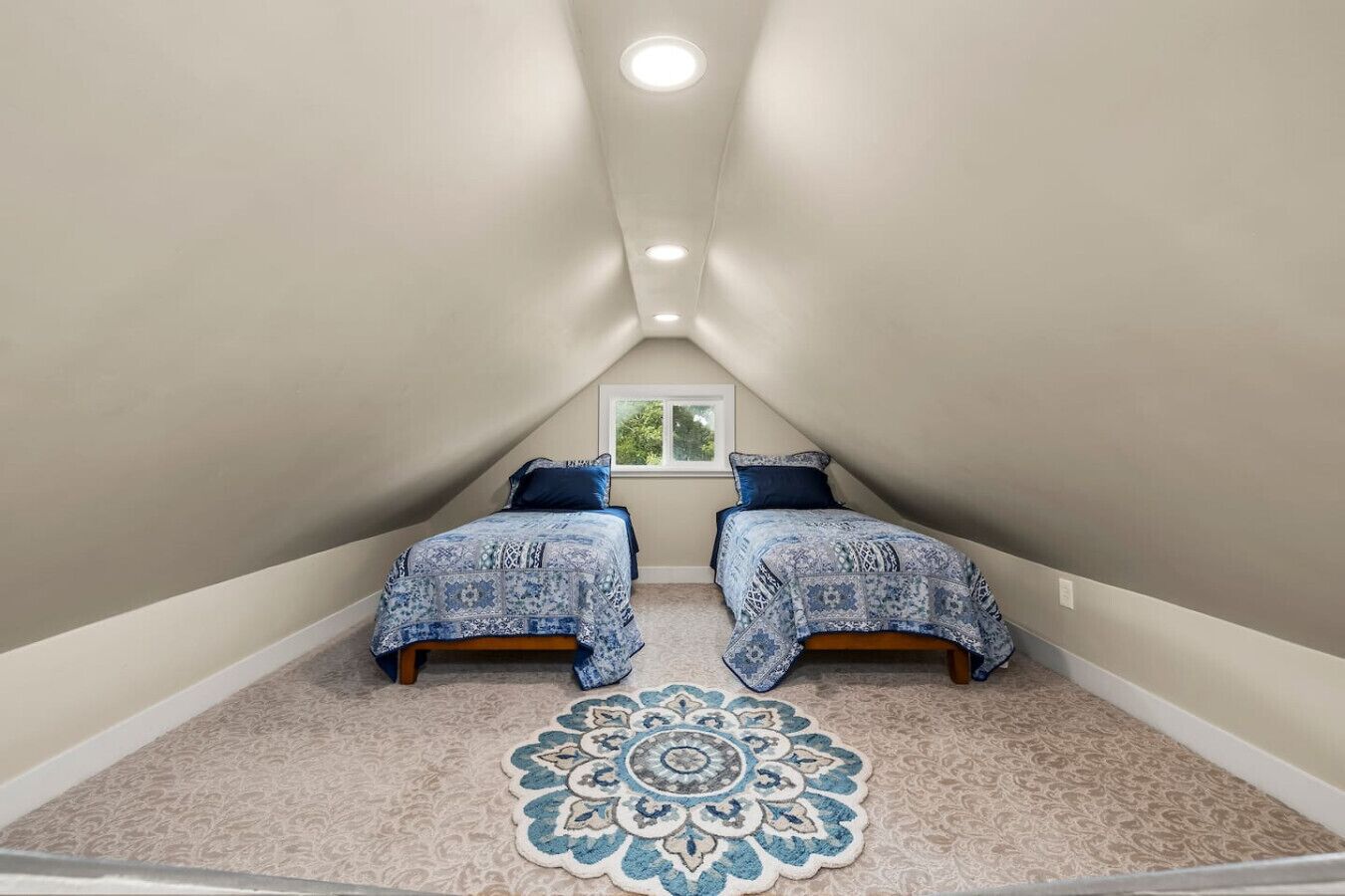 Een compacte zolderkamer met twee bedden, een groot decoratief vloerkleed en een raam met uitzicht.