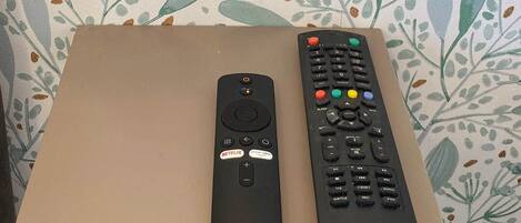 Tomada elétrica na parede com duas entradas USB, um controle remoto e um controle de TV sobre uma mesa, ao fundo papel de parede com motivos florais.