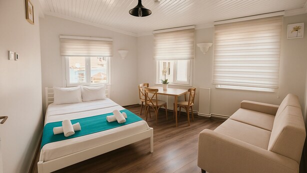A hotel room with a bed, a sofa, a dining table with chairs, and large windows with blinds.