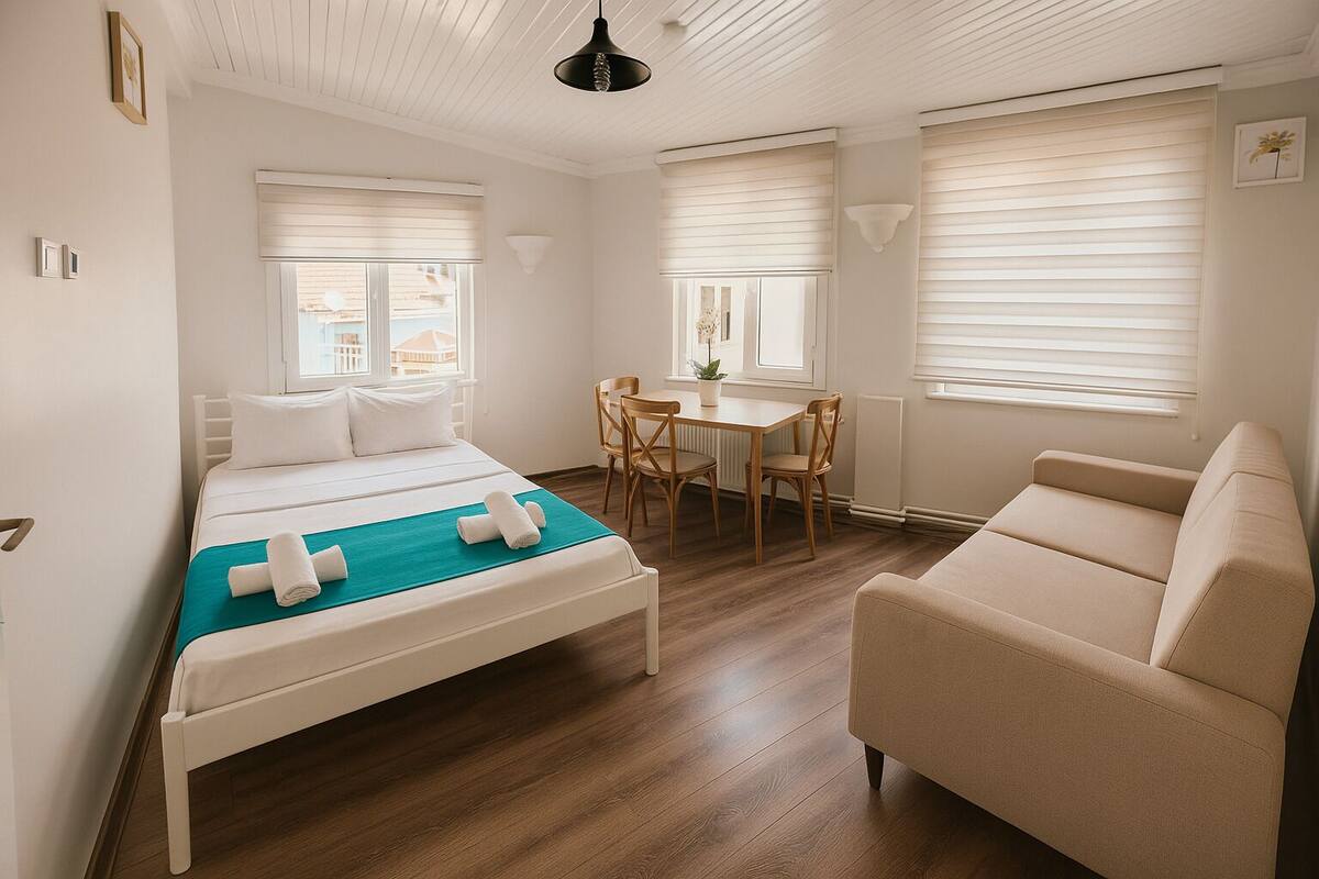 A hotel room with a bed, a sofa, a dining table with chairs, and large windows with blinds.