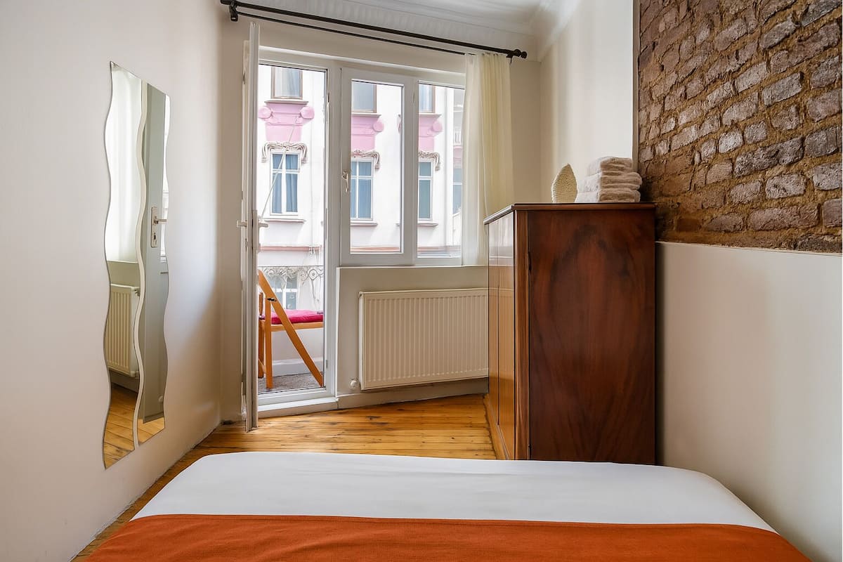 A bedroom with a wooden floor, a bed with an orange blanket, a wooden dresser, a mirror, and a view of a pink building through a glass door.