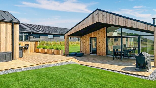 Une maison moderne avec une façade en bois, un patio couvert aménagé avec du mobilier d’extérieur et une grande porte vitrée donnant sur l’intérieur.