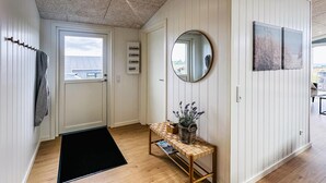 Un couloir doté d’un plancher en bois, d’un paillasson noir, d’un banc en bois avec un panier en osier, d’un miroir rond et d’une porte avec une fenêtre.