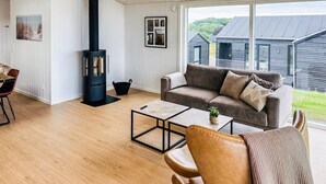 Un salon moderne avec un poêle à bois noir, un canapé gris, une table basse et une vue sur une maison, visible à travers de grandes fenêtres.