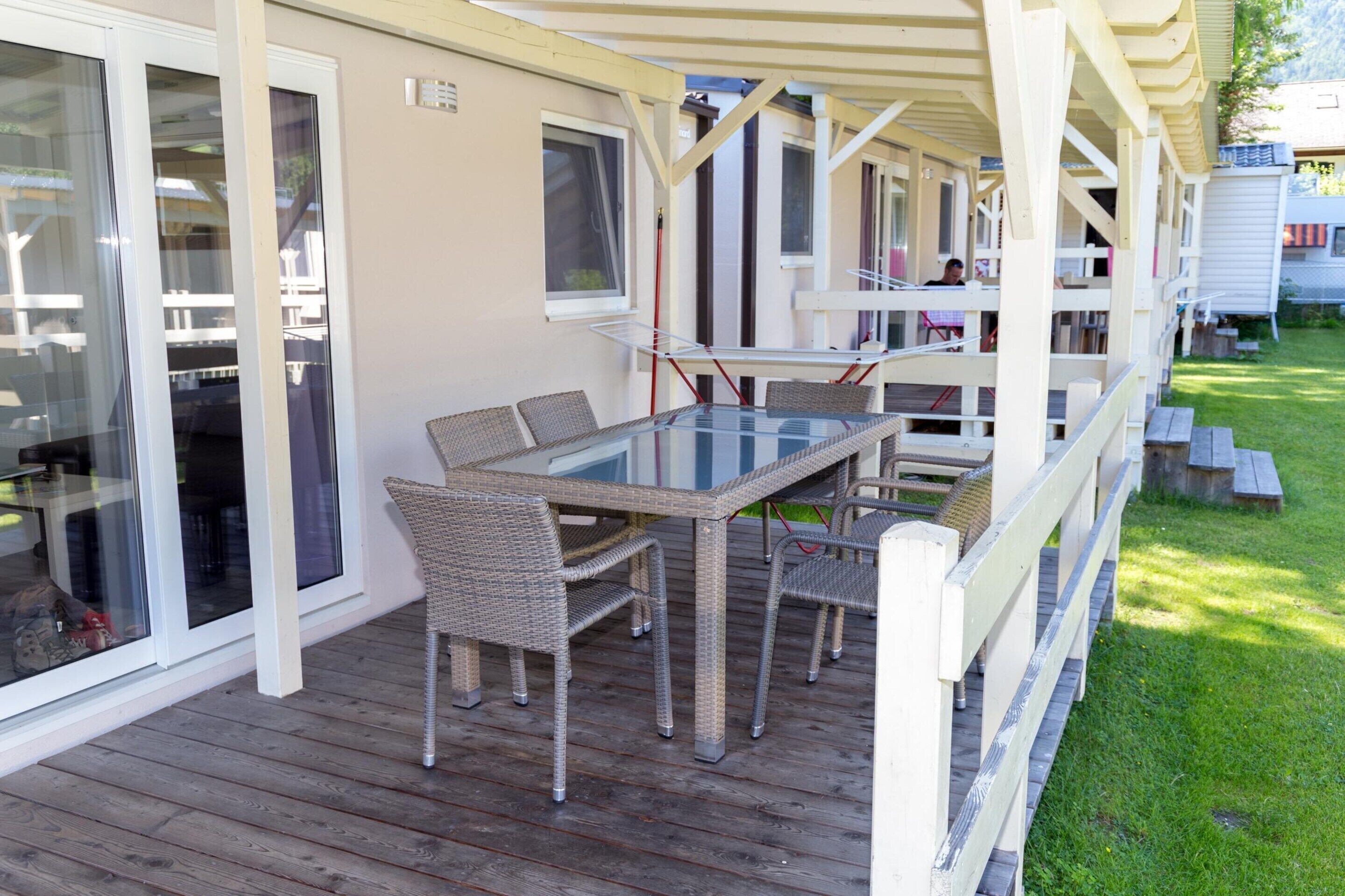 Eine überdachte Terrasse mit Glastisch und Korbstühlen, direkt neben einem Gebäude mit Glasbodenschiebetüren.