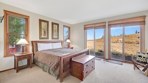 A bedroom with a large bed, two bedside tables with lamps, a wooden nightstand, a chair with a patterned cushion, and a view of a grassy landscape through large windows. - Snowcreek 811- Beautiful views, Spacious (Mammoth Lakes)