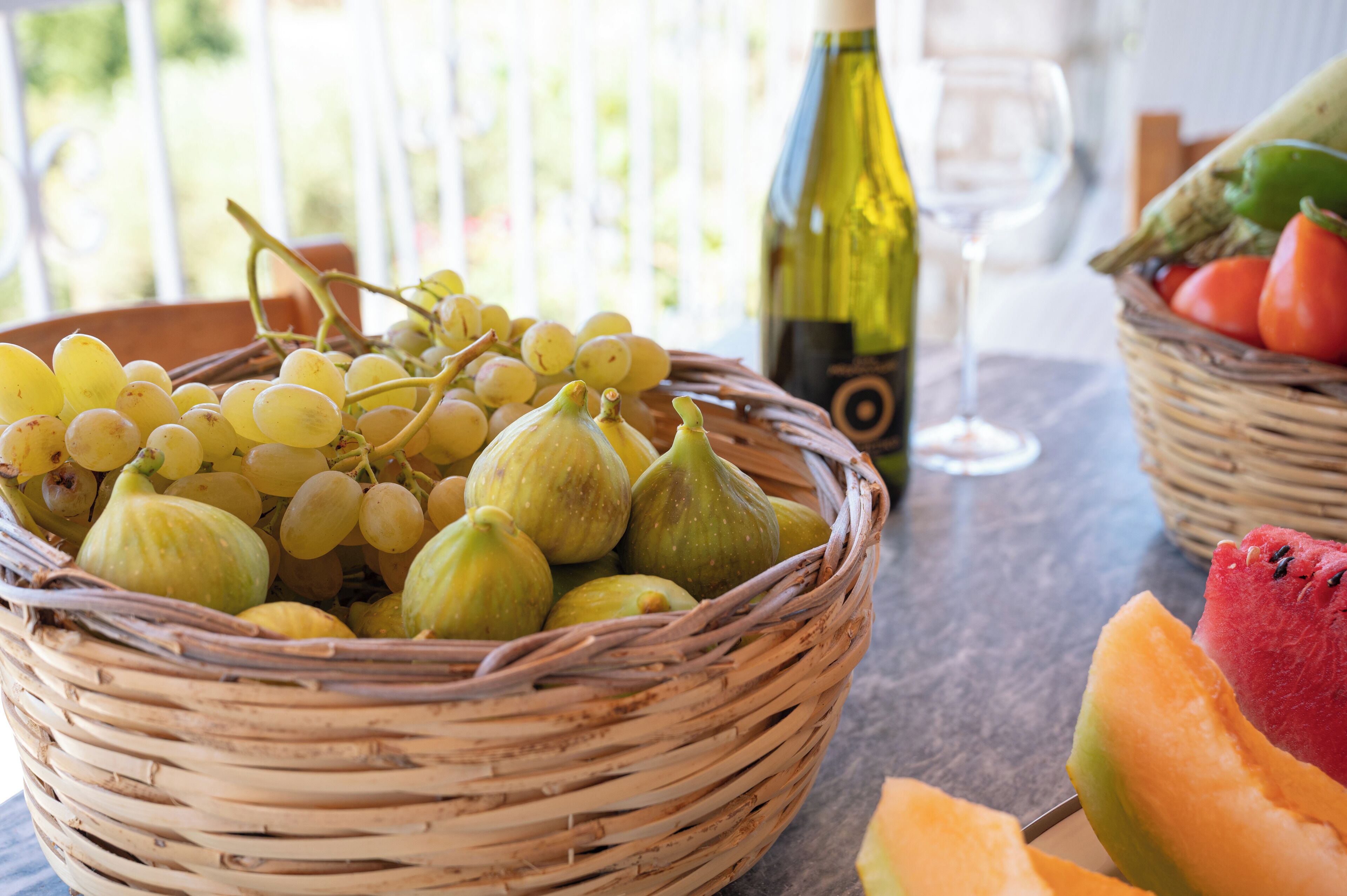 Una cesta de mimbre con uvas e higos, una botella de vino y una copa de vino sobre una mesa.