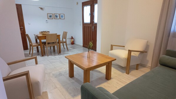 Un salón con una mesa de madera, dos sillas, un sofá y un sillón verde. Hay un extintor cerca de la puerta.