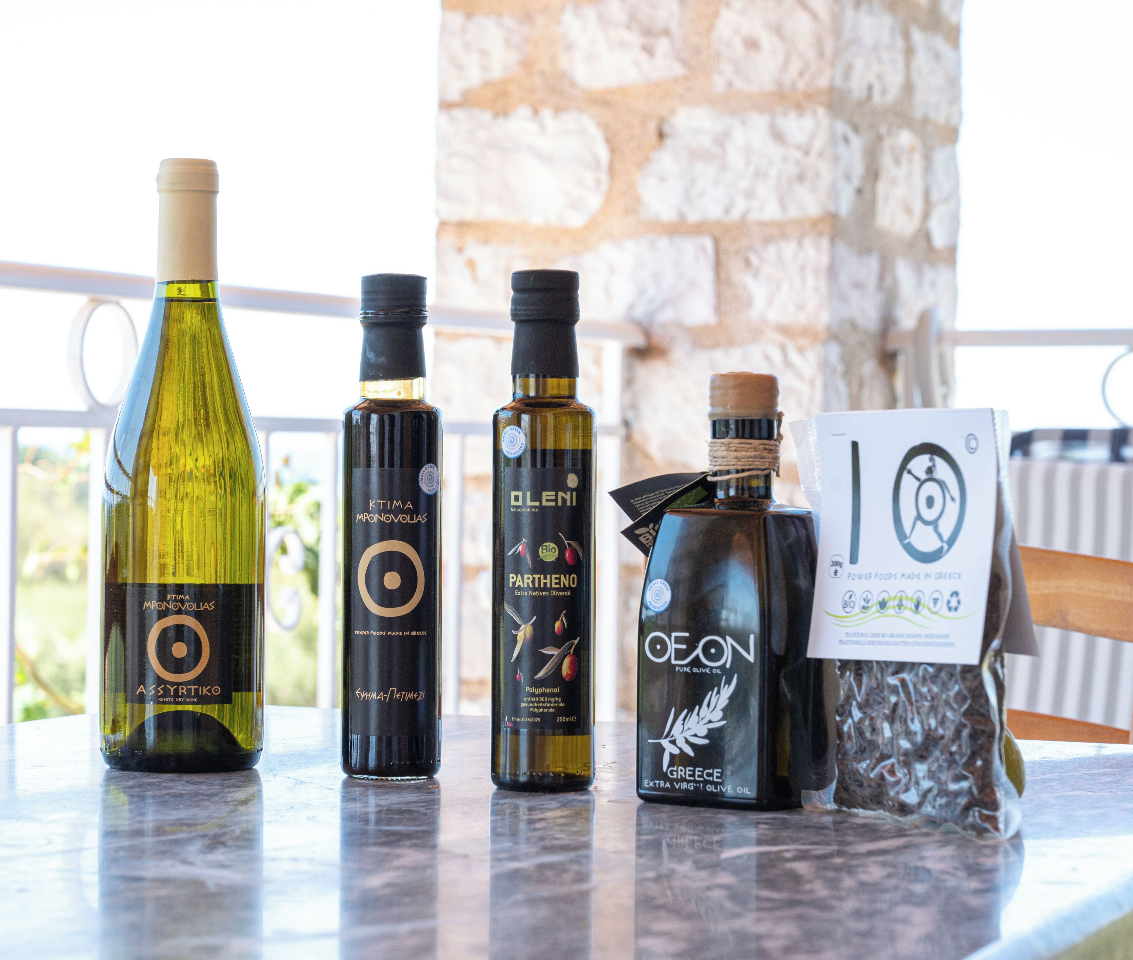 A selection of olive oil bottles and a bag of olives on a table with a stone wall in the background.