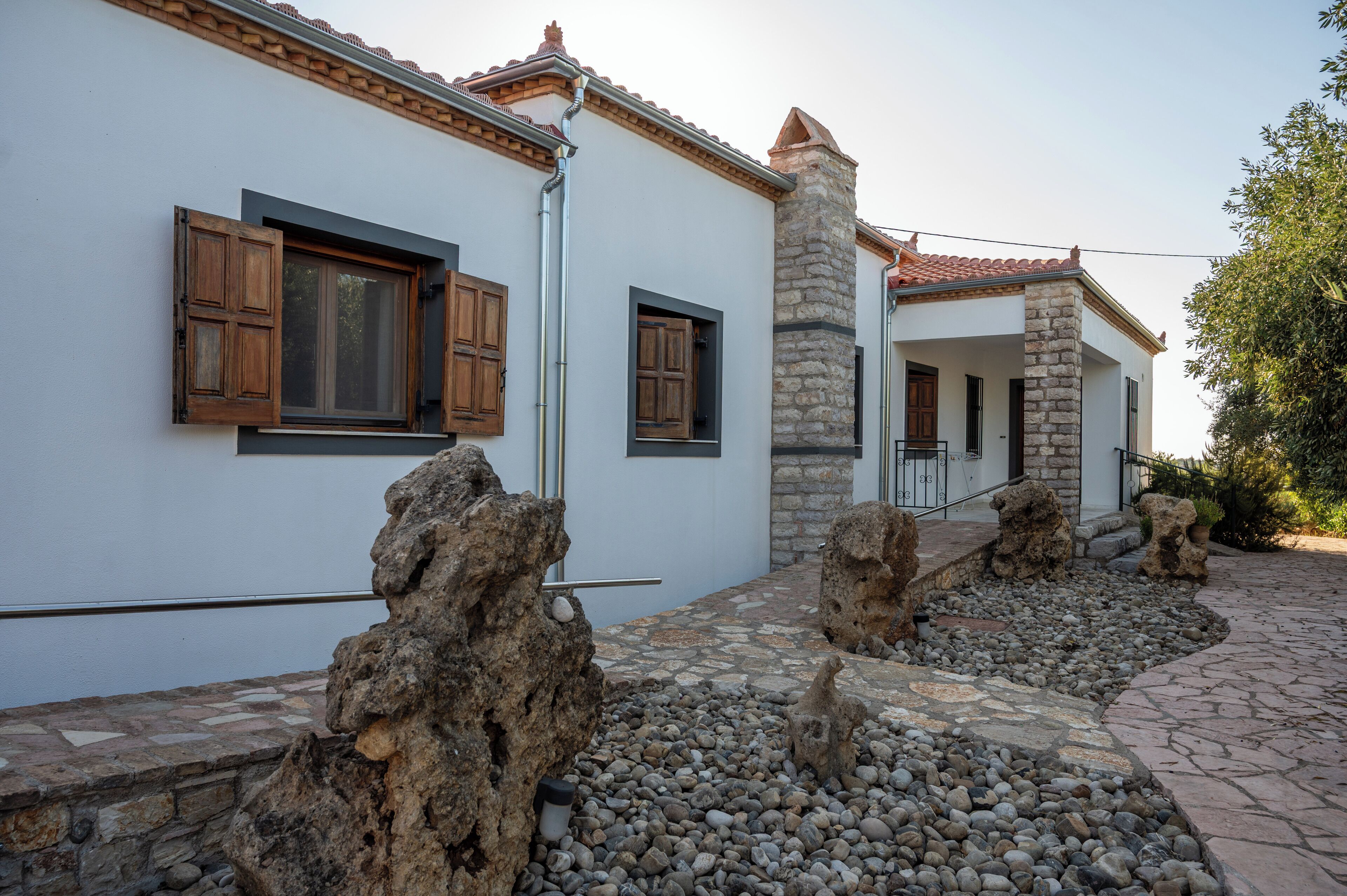 A building with white walls, stone pillars, and a tiled roof. There are wooden shutters on the windows and a stone pathway leading to the entrance.