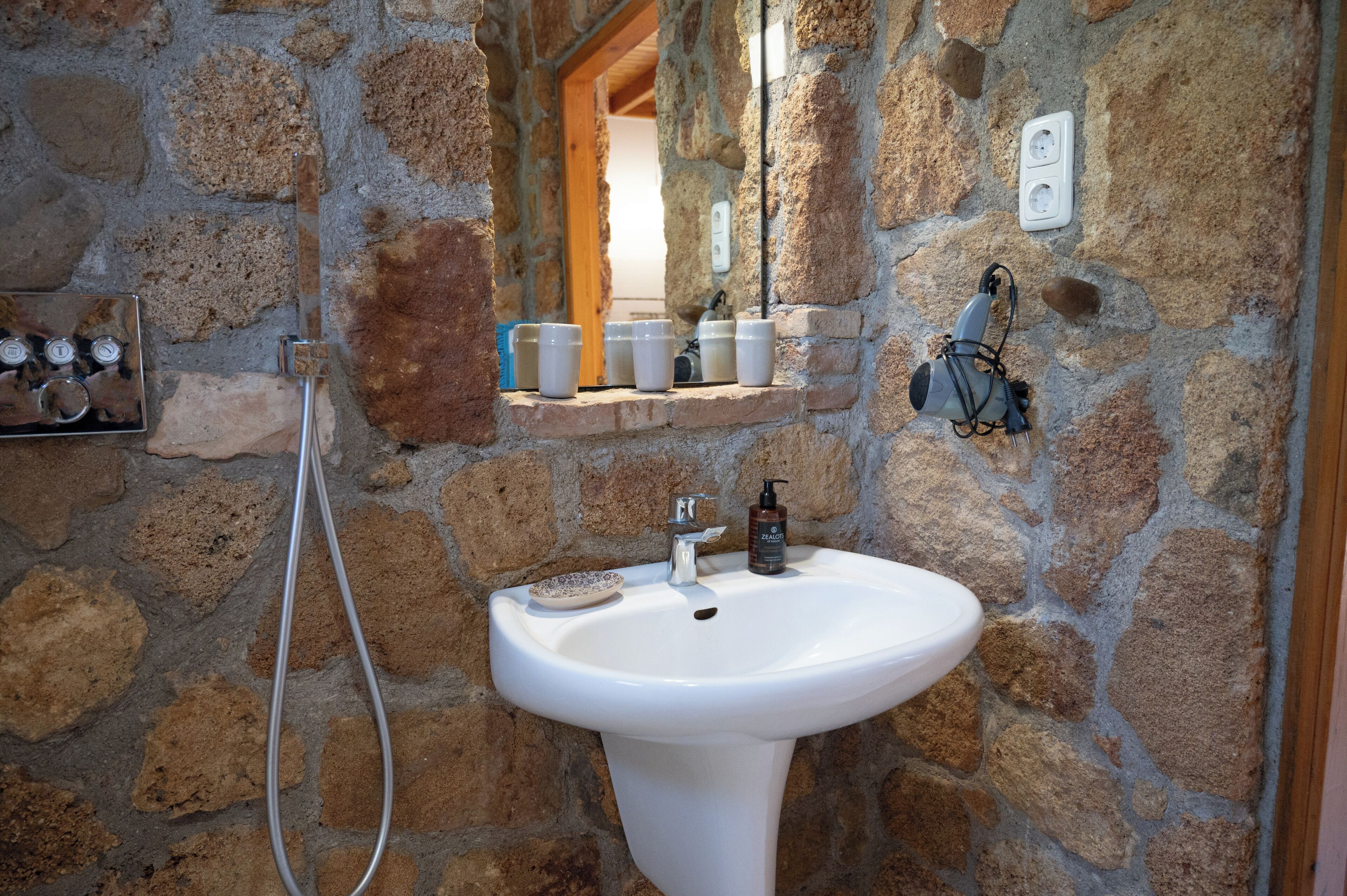 Un baño con paredes de piedra, un lavamanos blanco, un espejo y un estante con artículos de aseo.