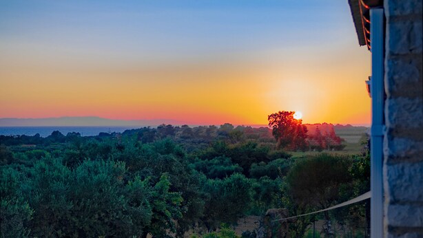 A sunset view from a balcony with a stone floor, potted plants, and a view of the surrounding landscape. - Terra Ionian (Pyrgos)