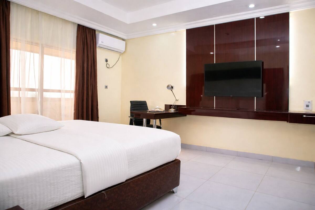 Quarto de hotel com cama, escrivaninha, cadeira, televisão fixada na parede e aparelho de ar condicionado.
